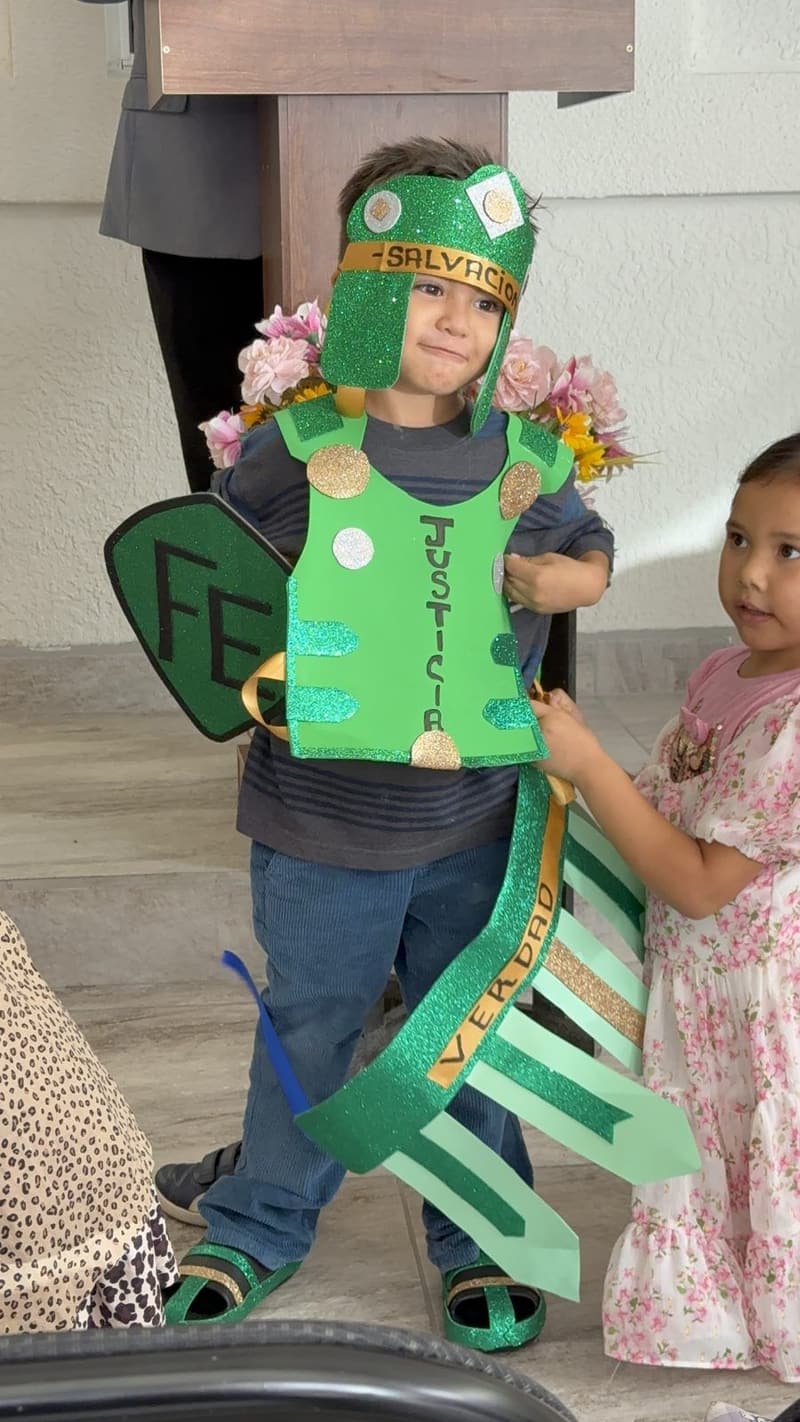 A boy wearing a handmade green helmet and breastplate of the armor of God, with a paper shield, while a girl stands beside him.