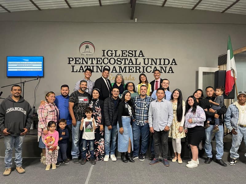 Members of the Ensenada church, adults and children, smiling together in front of the church name on the wall.