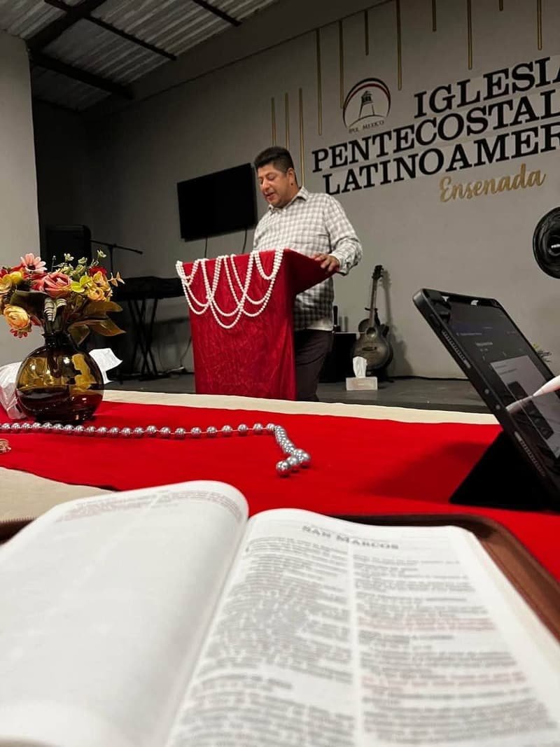 An open Bible in the foreground and a man speaking at a pulpit in the Ensenada church.