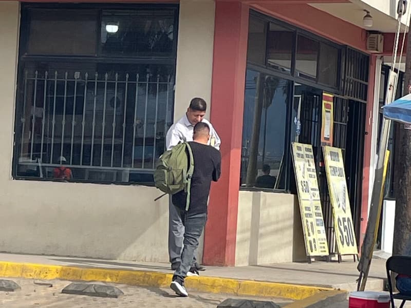 A church member talks with a man carrying a backpack outside a shop, seen from behind.